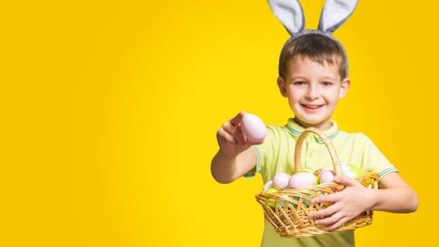 A happy smilling boy with Easter bunny ears holds a basket with painted eggs and shows one of them on a yellow background. Copy space. Easter day.