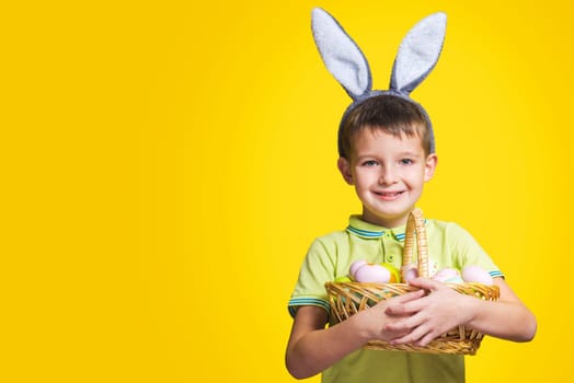 A happy smilling boy wearing Easter bunny ears holds a basket with painted eggs on a yellow background. Copy space. Easter day.