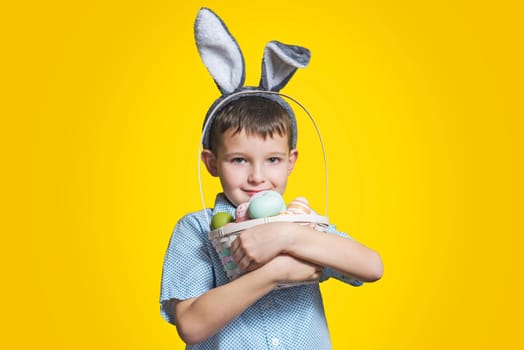 A happy smiling boy wearing Easter bunny ears holds a basket with painted eggs on a yellow background. Copy space. Easter day.