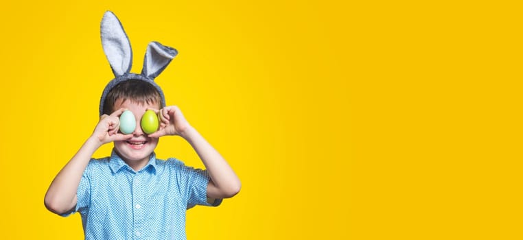 Cute boy with bunny ears keeping colored eggs near eyes on yellow background. Happy child boy with easter eggs and bunny ears on yellow background.