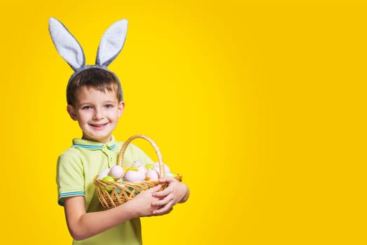 A happy smiling boy wearing Easter bunny ears holds a basket with painted eggs on a yellow background. Copy space. Easter day.