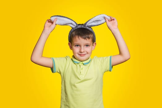 Little boy in t-shirt is touching his bunny ears and smiling while standing yellow background. Cute little child wearing bunny ears on Easter day.