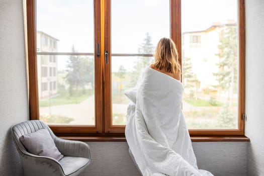 Beautiful young woman wrapped in a blanket standing near the window in the morning. Good morning, new day concepts.