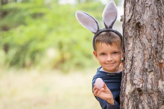 Cute little boy with rabbit ears holding easter egg on green natural background. Egg hunt at spring time. Happy Easter day.