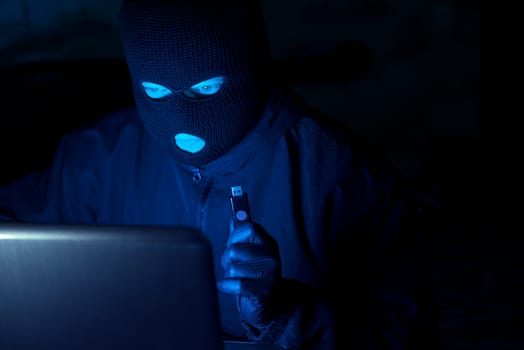 Cybercrime, hacking and technology crime. Masked hacker working in the darkness stealing data and personal identity information off a laptop computer.