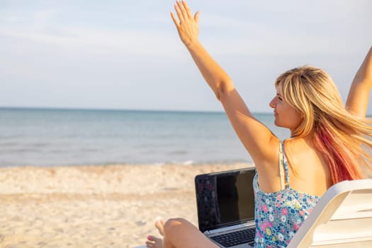 Rear view of young blonde woman, female freelancer working on laptop, keeping arms raised and celebrating success while sitting on the tropical sandy beach.