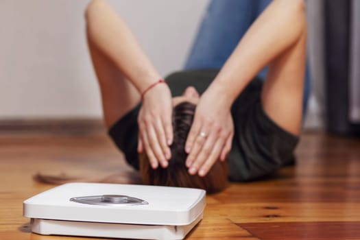 Scales and depressed young woman lying on the floor holding her head and covering her face with her hands.