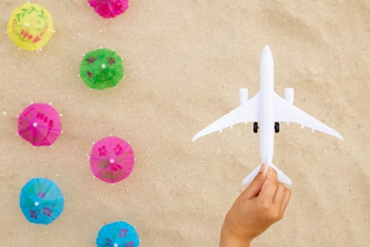 Childs hand with toy airplane under over a yellow sandy beach with beach umbrellas. Trip journey and holiday summer concepts