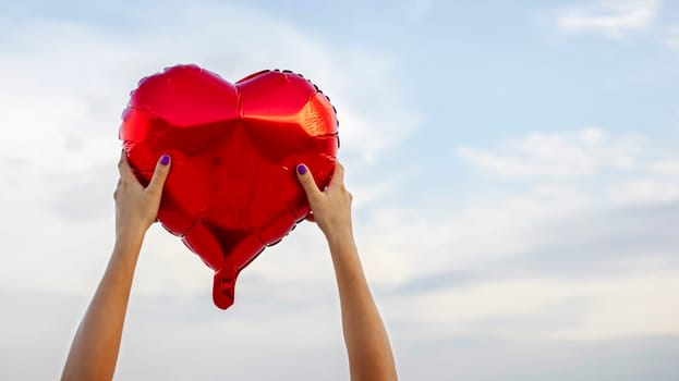 Womans hands with heart shaped balloon on background of sky. Valentine day and romance concept.
