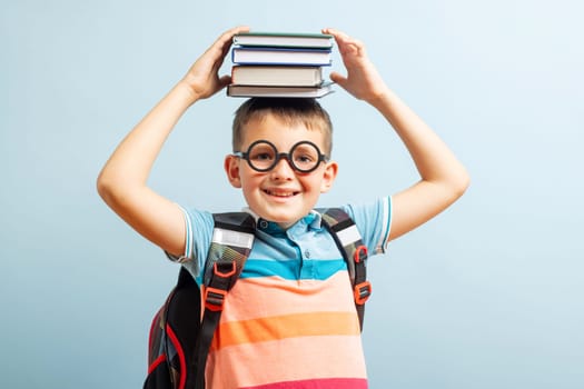 Funny smiling school boy in glasses holds books on his head on blue background. Back to school.