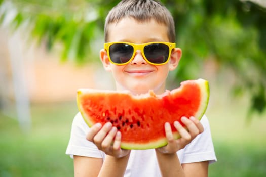 Cute boy in yellow sunglasses eating watermelon outdoors in summer. Healthy eating seasonal berries and fruits.