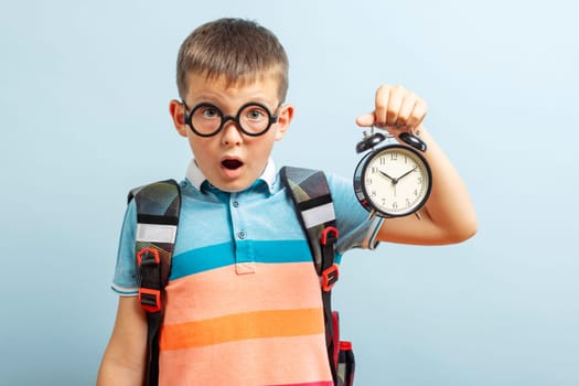Amazed child in glasses listen alarm clock ringing showing time, amazement. Schoolchild with clock alarm on blue background.