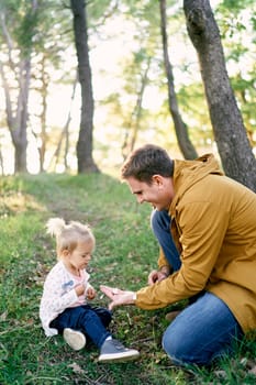 Dad holds out a tiny mushroom in the palm to a little girl sitting in the forest on the grass. High quality photo