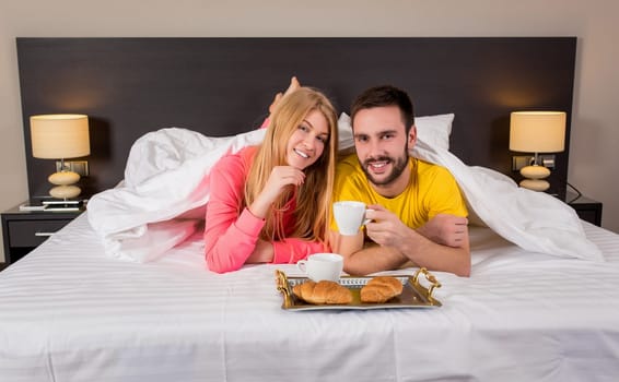 Happy young couple having breakfast tray on bed at home in bedroom. Young couple drink coffee in bed, happy smile woman man lovers