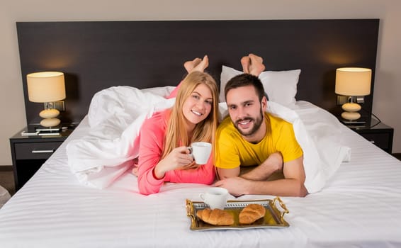 Happy young couple having breakfast tray on bed at home in bedroom. Young couple drink coffee in bed, happy smile woman man lovers