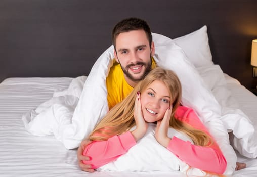 Young lovely couple lying and have fun in a bed, happy smile looking at camera, cover under blanket. Family, bedtime and happiness concept