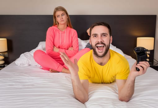 Young couple having playing videogames in bed. Man playing video game, upset woman on background.