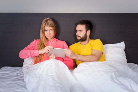 Young sweet couple at bed with Tablet Gadget, man takes it girl. Concept about technology and people