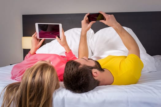 Young couple lying on their backs on the bed, the husband with a mobile phone and the wife with tablet