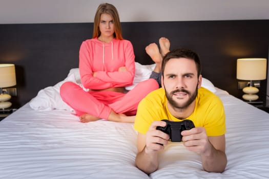 Young couple having playing videogames in bed. Man playing video game, upset woman on background.