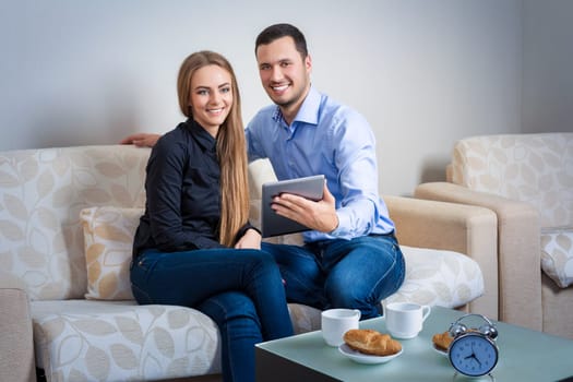 Beautiful laughing young couple, drinking coffee with croissants, sitting on a sofa, with electronic tablet and looking at the camera.