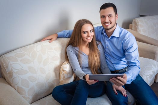 Beautiful laughing young couple, sitting on a sofa, with electronic tablet and looking at the camera.