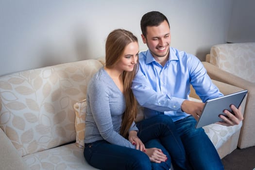 Beautiful young couple laughing, sitting on sofa, sharing photos or other information, displayed on an electronic tablet with each other