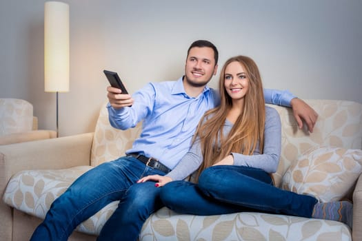 Happy couple sitting on sofa with remote control in hands and watching television together