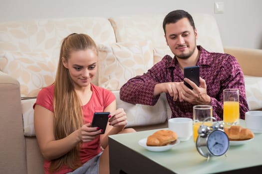Cute young couple on the floor beside the couch having breakfast, eating croissants, drinking orange juice and looking at mobile phone