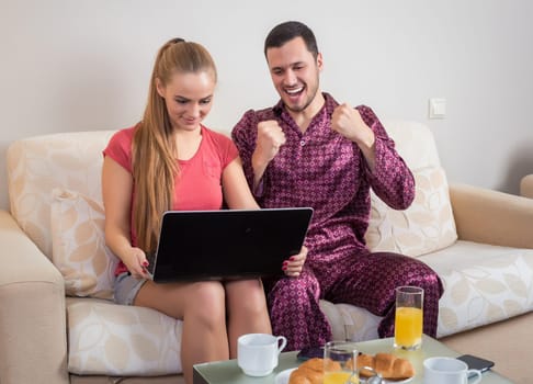 Cute young couple on the couch having breakfast, eating croissants, drinking orange juice in front of a laptop computer