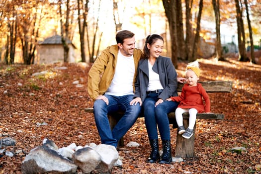 Smiling mom and dad look at a little girl sitting with her on a bench in the autumn forest. High quality photo