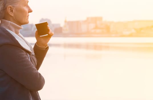 An adult beautiful woman holds a glass of aromatic coffee in her hands and enjoys the taste and atmosphere at sunset, views of the city and the river.