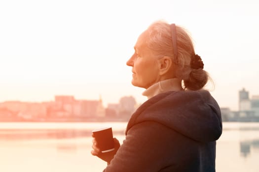 An adult beautiful woman holds a glass of aromatic coffee in her hands and enjoys the taste and atmosphere at sunset, views of the city and the river.