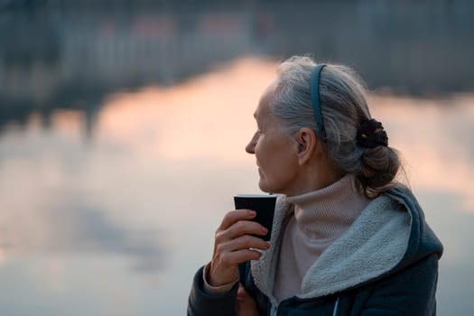 An adult beautiful woman holds a glass of aromatic coffee in her hands and enjoys the taste and atmosphere at sunset, views of the city and the river.