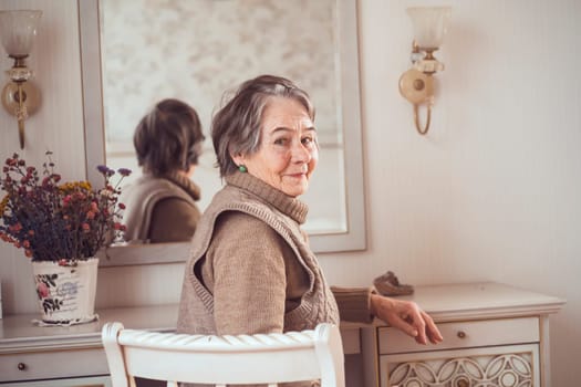 An elderly smiling woman of 80+ years of age spends a good time at home, a grandmother takes care of her appearance, looks in the mirror, sits at a dressing table in her bedroom.