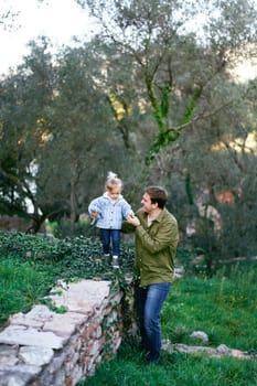 Little girl walks along a stone fence, supported by her father by the hand. High quality photo