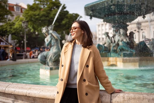 Woman wear glasses and stylish autumn coat sitting on fountain smiling looking away. Young adult girl resting on fountain Rossio square in Lisbon. Happy female relaxing in fresh air