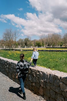 Little girl walks along a stone fence holding her mother hand. Back view. High quality photo