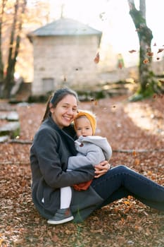 Smiling mom hugs a little girl on her lap sitting on the ground in an autumn park. High quality photo