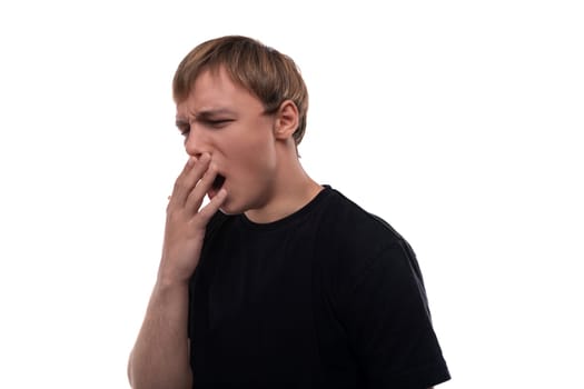 A handsome young man with blond hair dressed in a black T-shirt is tired and yawns.