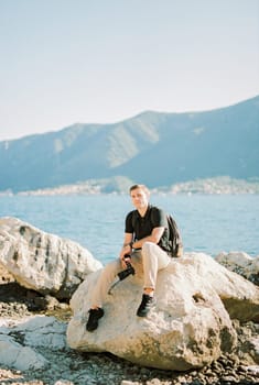 Photographer with a camera in his hand sits on a large boulder by the sea. High quality photo