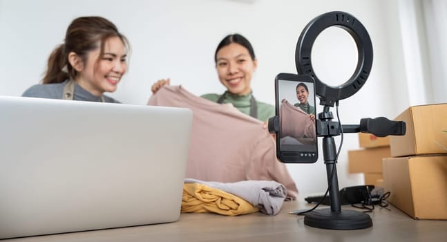 Two asian woman business owner at store showing clothes in front of smartphone to recording vlog video live streaming for sale fashion clothing to customer, Online shopping concept.
