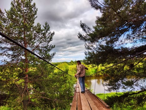 A girl on an old wooden suspension bridge among spruce or pine trees in nature. A tourist on a trip and landscape with trees on a cloudy autumn or spring day