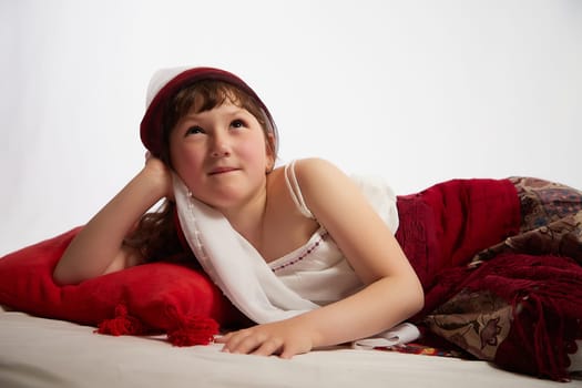 Portrait of Little girl in a stylized Tatar national costume having rest with a red pillow on a white background in the studio. Photo shoot of funny young teenager who is not a professional model