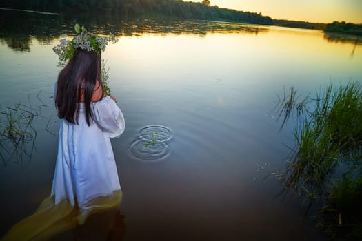 Adult mature brunette woman in a white dress, sundress and a wreath of flowers in summer in water of river or lake in evening at sunset. Celebration of the Slavic pagan holiday of Ivan Kupala