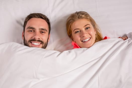 Young lovely couple lying and have fun in a bed, happy smile looking at camera, cover under blanket. Family, bedtime and happiness concept