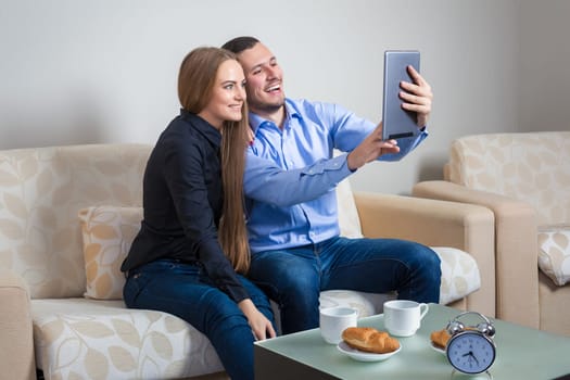 Beautiful young couple laughing, bvsitting on a sofa, sharing photos or other information, displayed on an electronic tablet with each other and drinking coffee with croissants