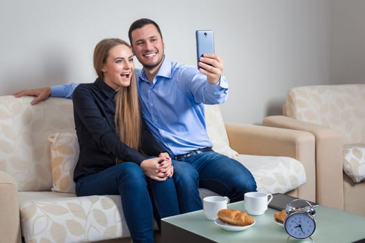 Beautiful young man and woman doing selfie with telephone camera, happy people taking picture smiling to the camera, bvsitting on a sofa