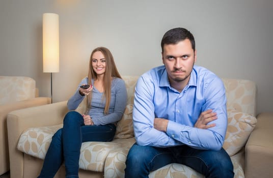 Portrait of couple sitting on sofa watching television. Image of woman with remote control in hands and upset men