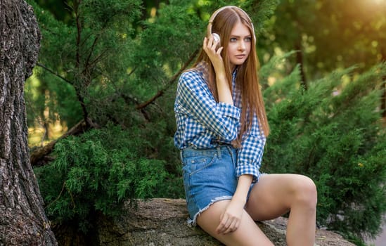 Young woman cute brunette girl sitting on stone on headphones, sun light flares of rays, looks into the distance, on summer outdoor copy space background